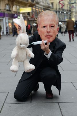 Als Minister verkleidete Aktivistin, die ein Stoffkaninchen mit einer Spritze bearbeitet.